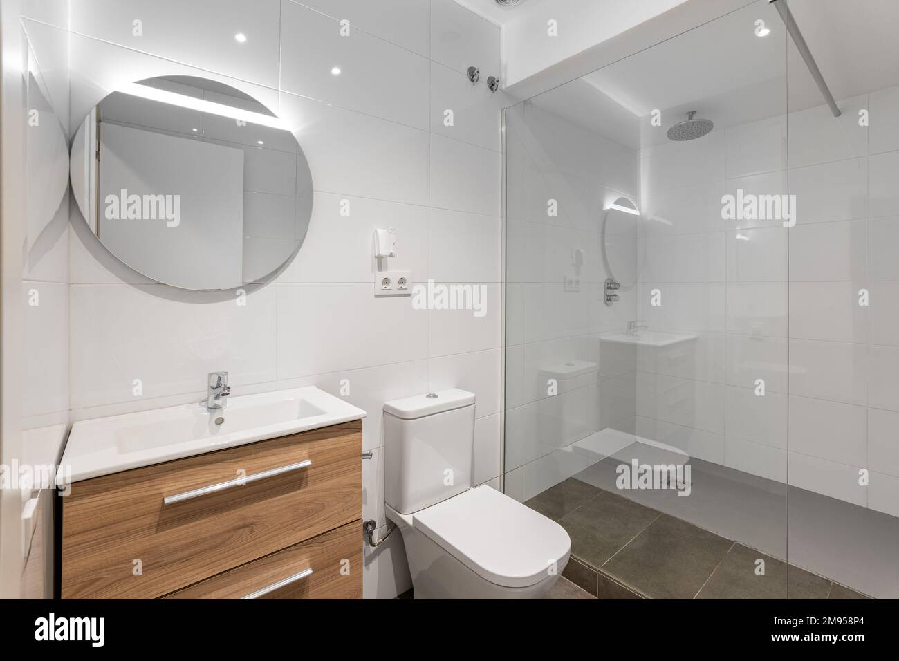 Bathroom with a toilet bowl and a sink made of white durable ceramics and wooder drawers. Shower with glass wall. Larger round mirror on the wall Stock Photo