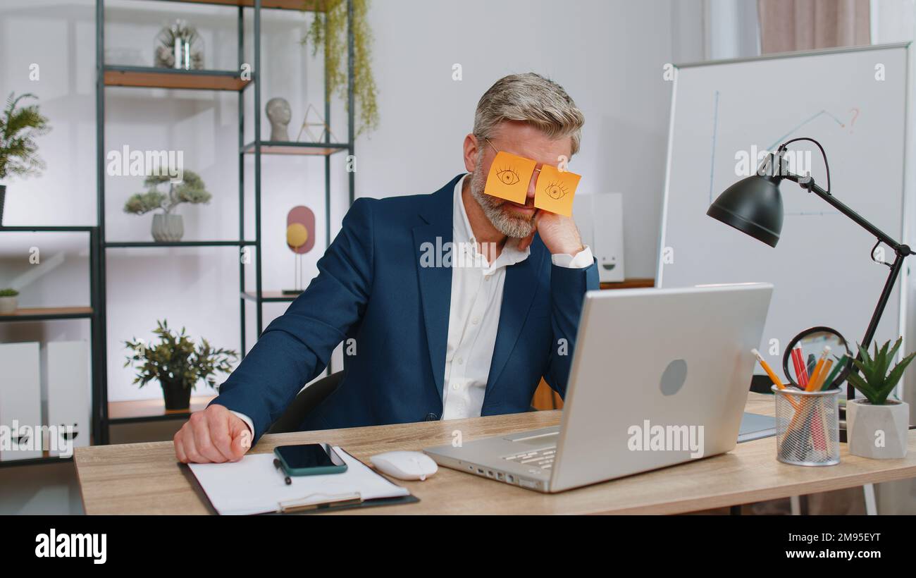 Young business man with fake eyes painted on paper stickers sleeping at  workplace in office Stock Photo
