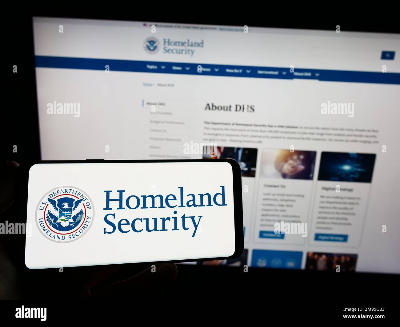 Person holding cellphone with seal of US Department of Homeland Security (DHS) on screen in front of webpage. Focus on phone display. Stock Photo