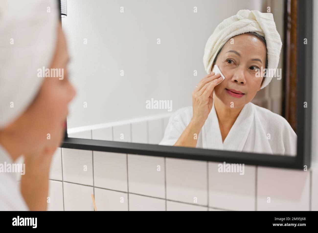 Beautiful and attractive mature aged Asian lady looking in the mirror, applying anti-aging skincare treatments with cotton pad after shower or removin Stock Photo
