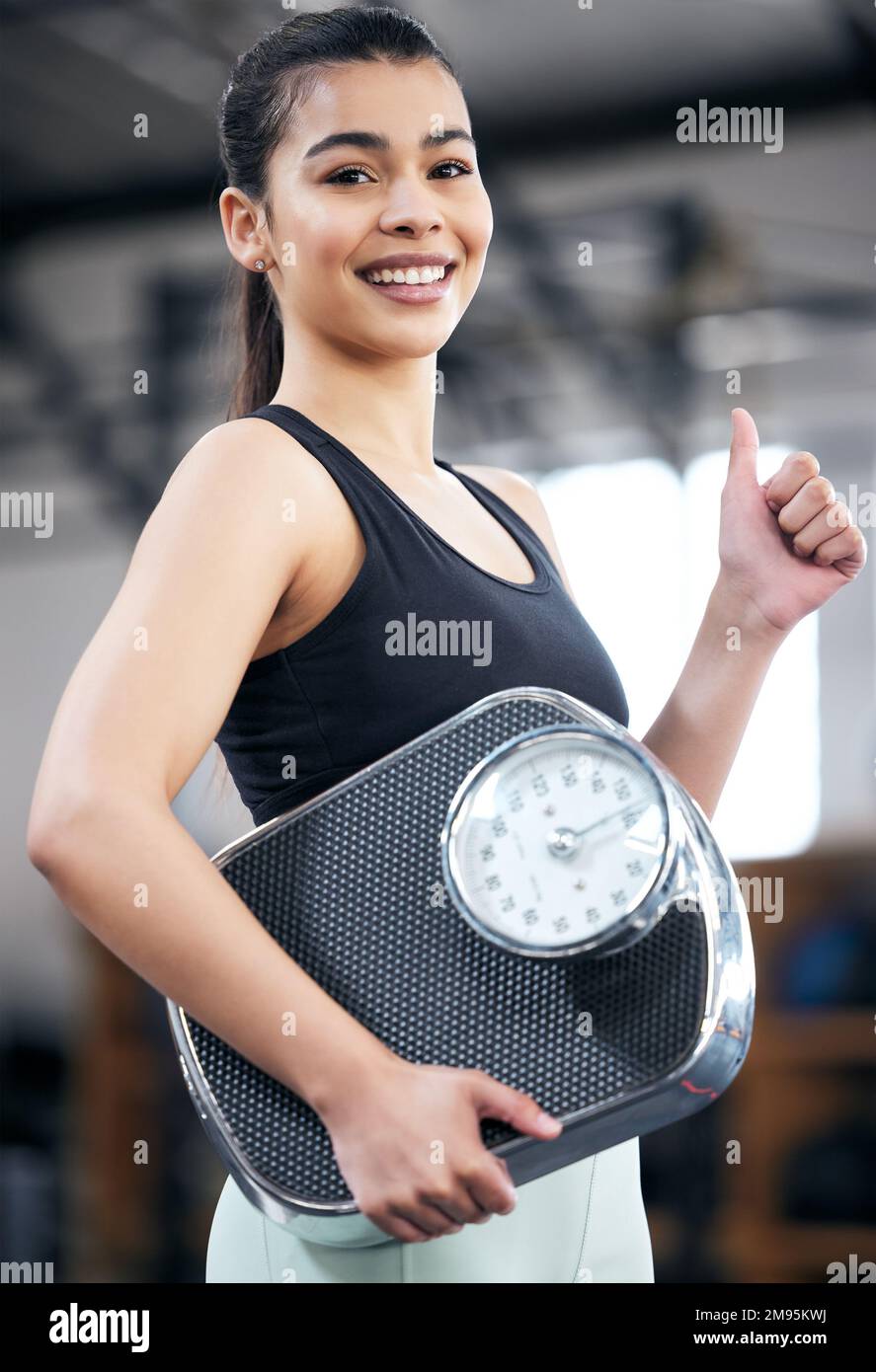 Im well on my way to reaching my goal weight. Portrait of a fit young woman holding a scale and showing thumbs up in a gym. Stock Photo