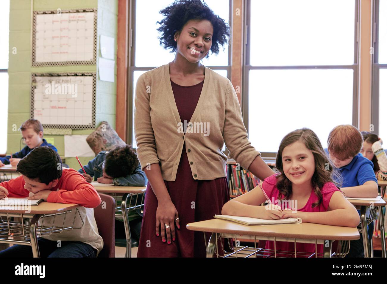 Education, learning and portrait of teacher with girl writing exam or test at Montessori school with students. Black woman, happy children at desk and Stock Photo