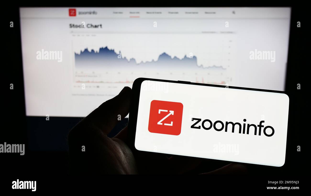 Person holding mobile phone with logo of US software company ZoomInfo Technologies Inc. on screen in front of web page. Focus on phone display. Stock Photo