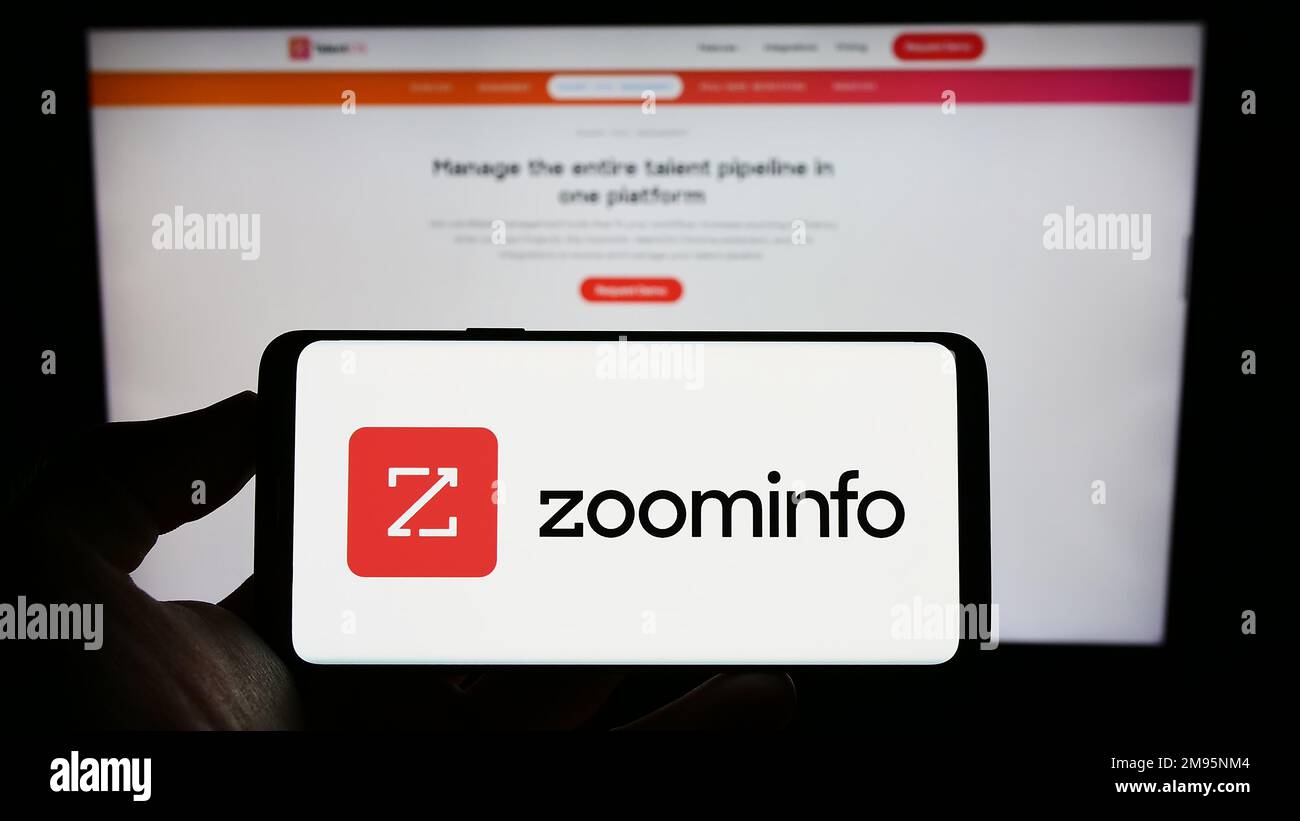 Person holding cellphone with logo of American software company ZoomInfo Technologies Inc. on screen in front of webpage. Focus on phone display. Stock Photo
