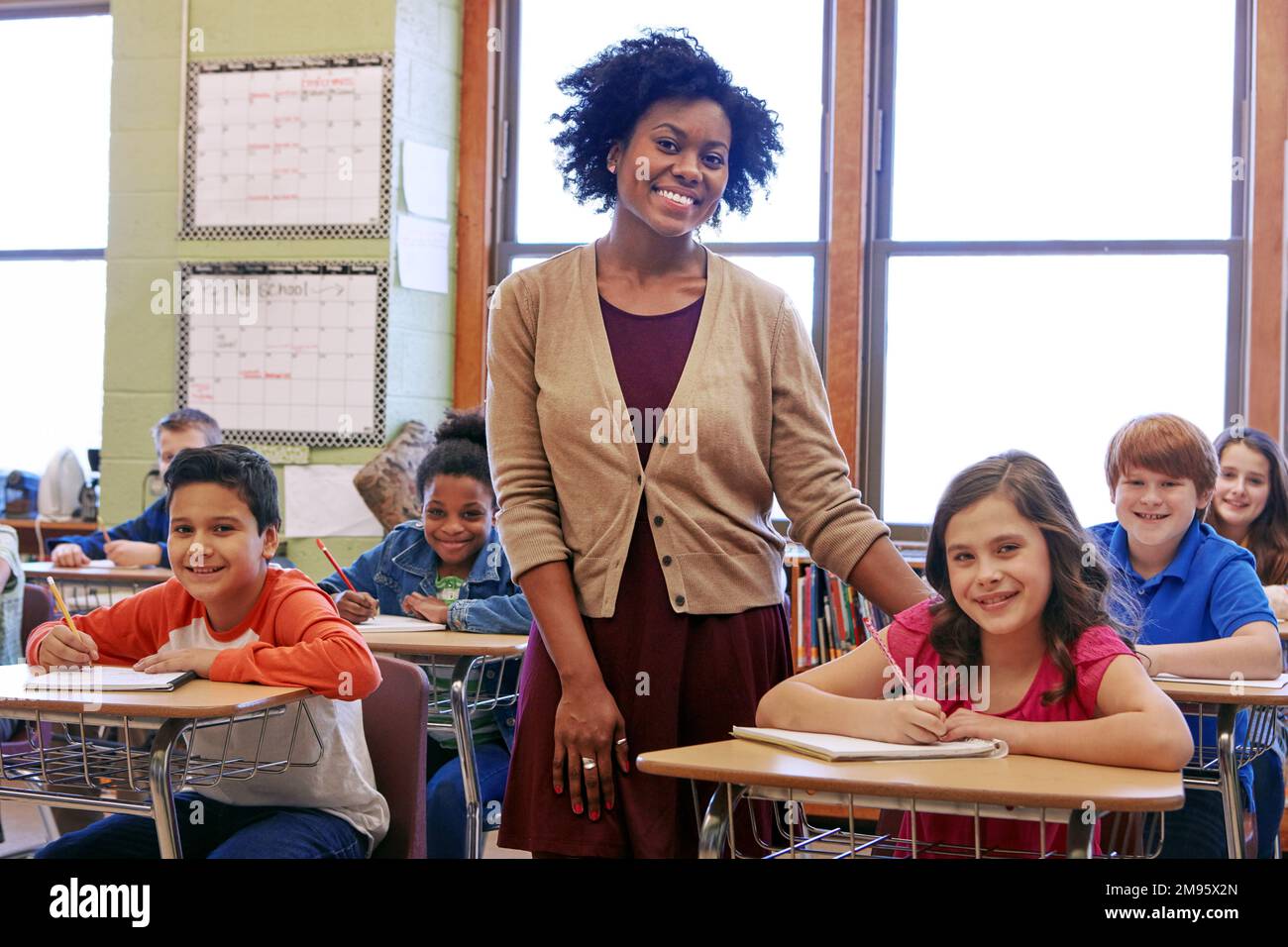 Education, learning and teacher in classroom with kids writing exam or test at Montessori school. Portrait of black woman, happy children at desk and Stock Photo