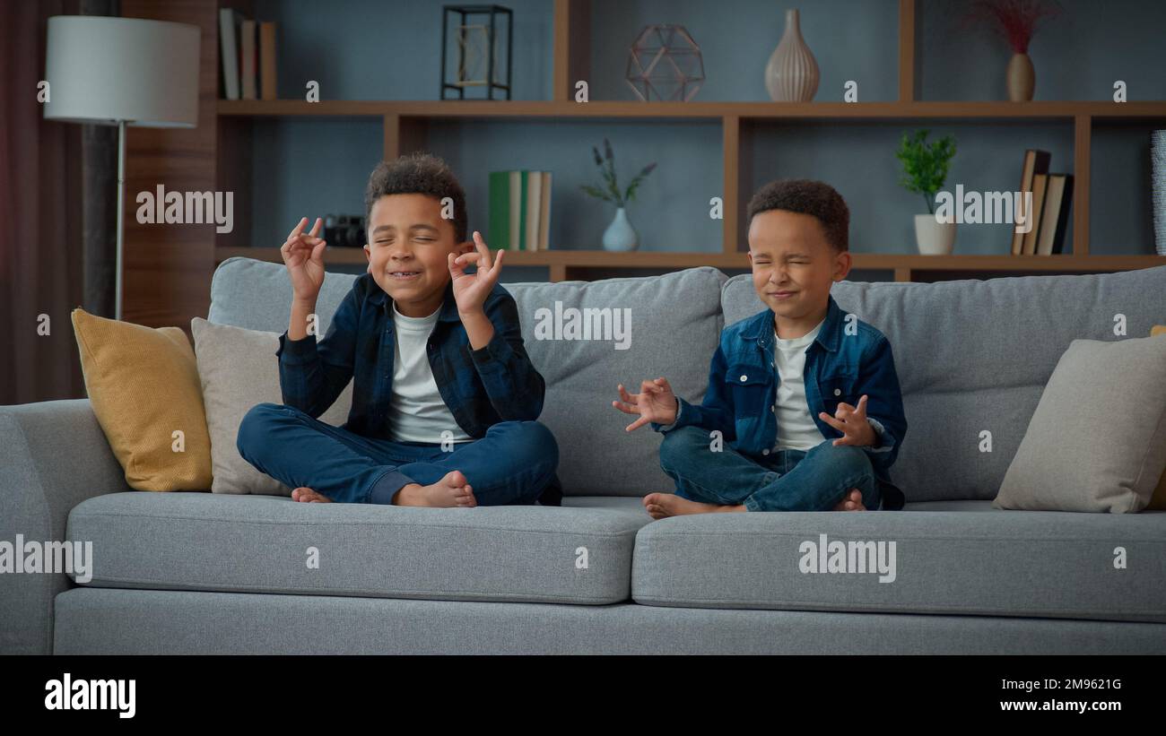 Two funny little African American ethnic children boys brothers siblings kids meditating together sitting on couch in lotus position. Small schoolboys Stock Photo