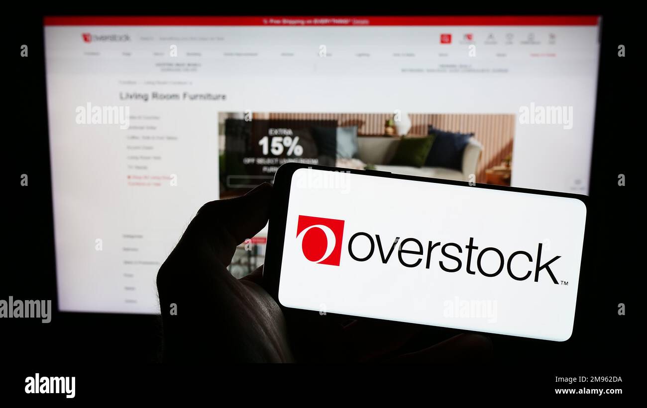 Person holding cellphone with logo of US e-commerce company Overstock.com Inc. on screen in front of business webpage. Focus on phone display. Stock Photo