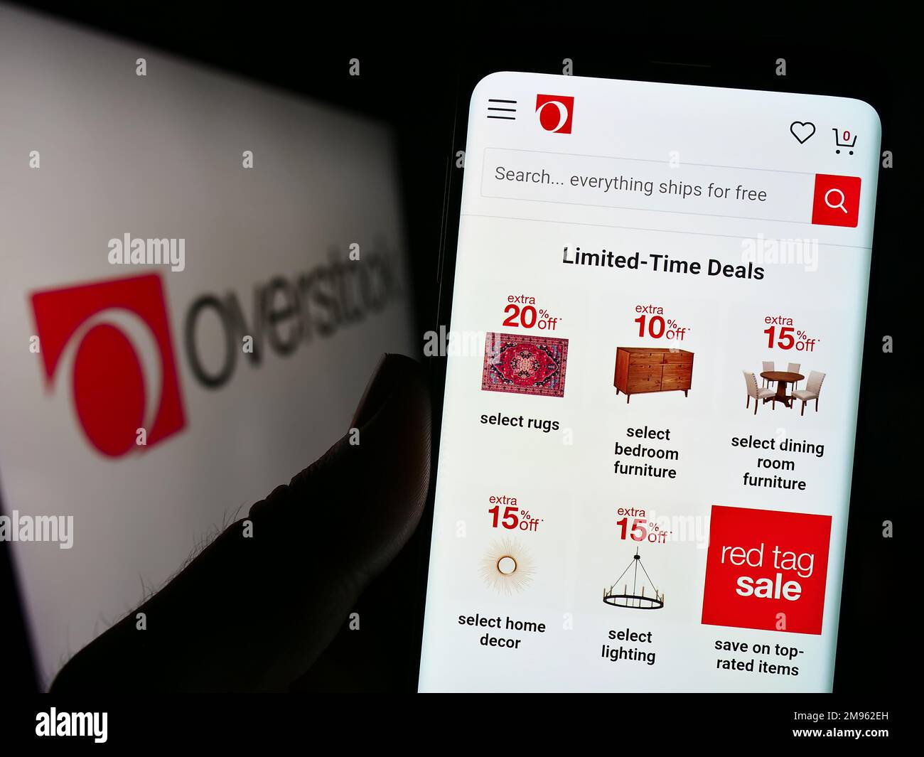 Person holding smartphone with website of US e-commerce company Overstock.com Inc. on screen in front of logo. Focus on center of phone display. Stock Photo