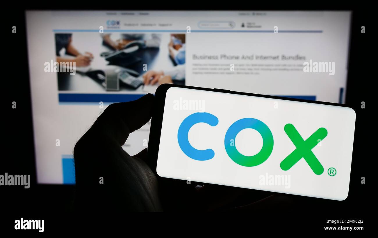 Person holding mobile phone with logo of US cable TV company Cox Communications Inc. on screen in front of web page. Focus on phone display. Stock Photo