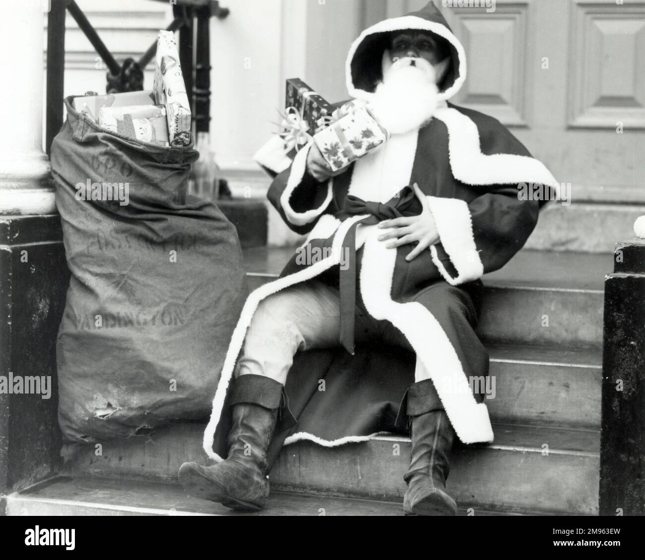 Father Christmas sitting on some steps in a street with a sackful of presents. Stock Photo