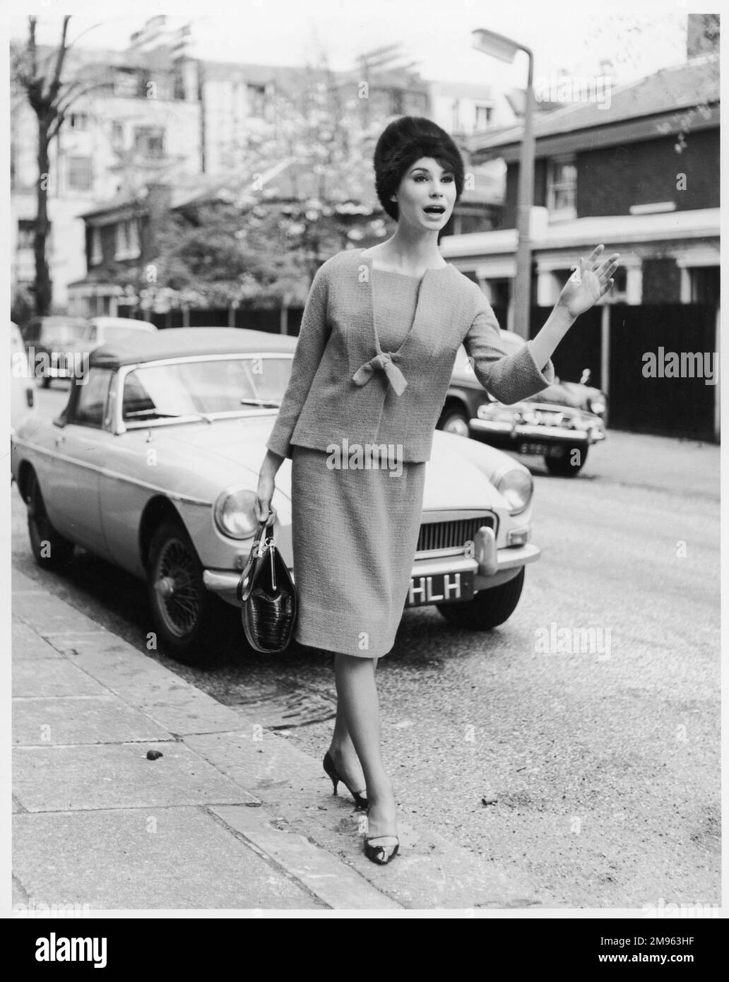 A woman in smart stylish suit of straight knee-length skirt & a collarless sac like short jacket with tie fastening, fur hat & stiletto heels hails a taxi. Stock Photo