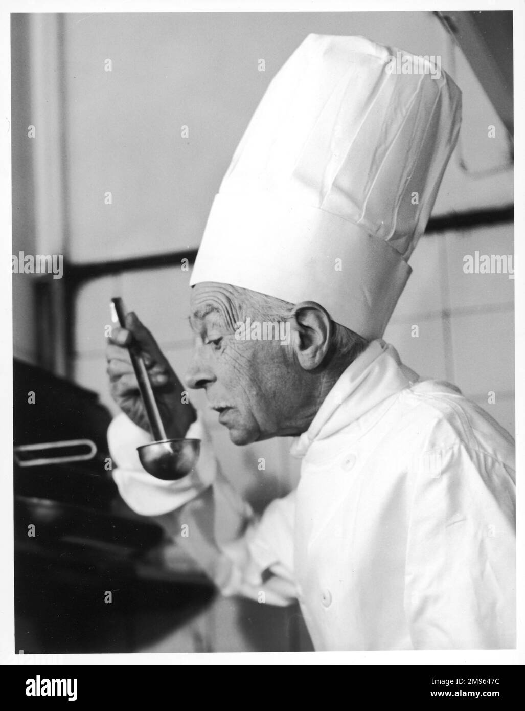 A chef in a chef's hat tastes some soup or other food from a ladle. Stock Photo