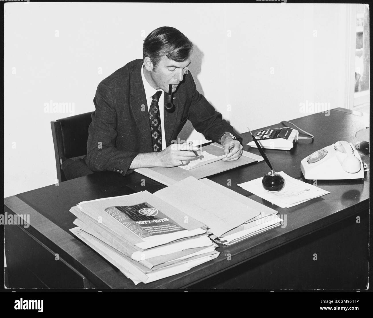 'The Boss' smoking a pipe and reading a letter, piles of files, a telephone and calculator on his desk. Stock Photo