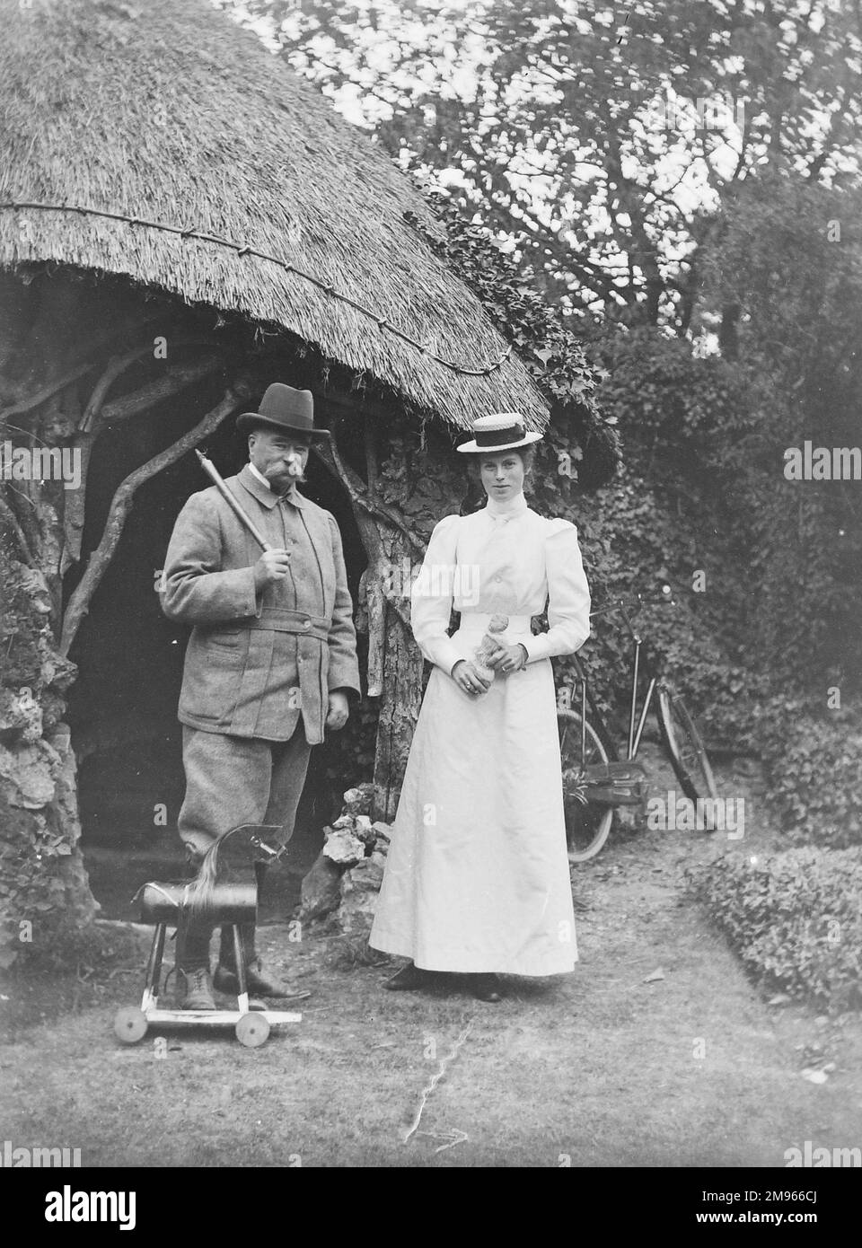 A moustachioed country gentleman and younger lady (his daughter?) in white dress and boater stand close to a thatch-roofed grotto and a child's hobby-horse Stock Photo