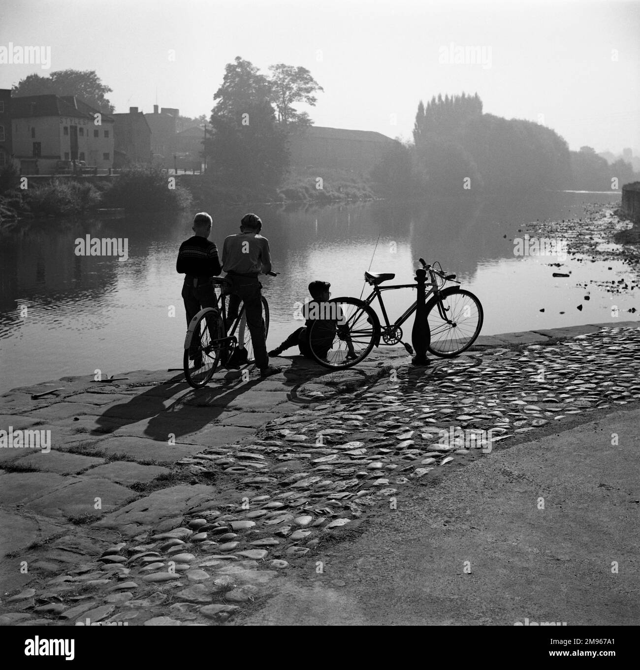 Three boys with their bikes admire the still river in the clear evening light at Bewdley Bridge, Worcestershire. Photograph by Norman Synge Waller Budd Stock Photo