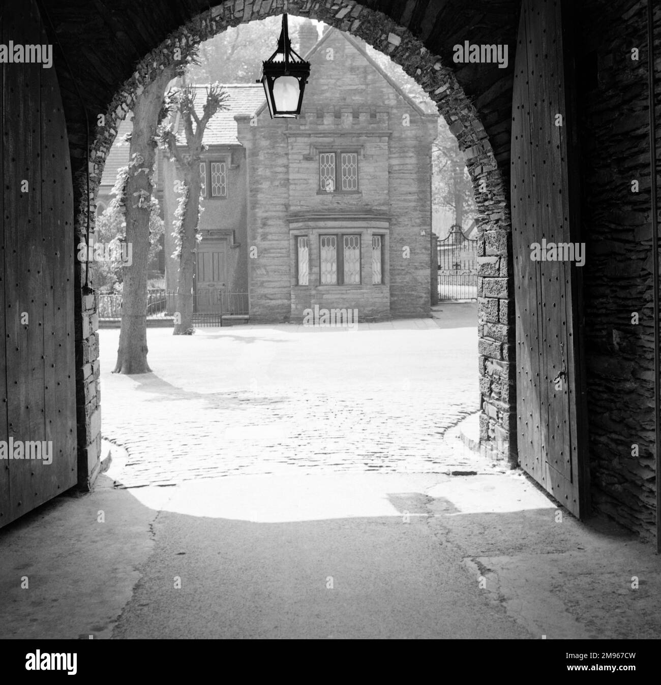 An open gateway with a fine gas lamp hanging at the point of the arch and two large wooden doors both opened fully. The open portal reveals a view though to an attractive small stone house in the sunshine. Photograph by Norman Synge Waller Budd Stock Photo