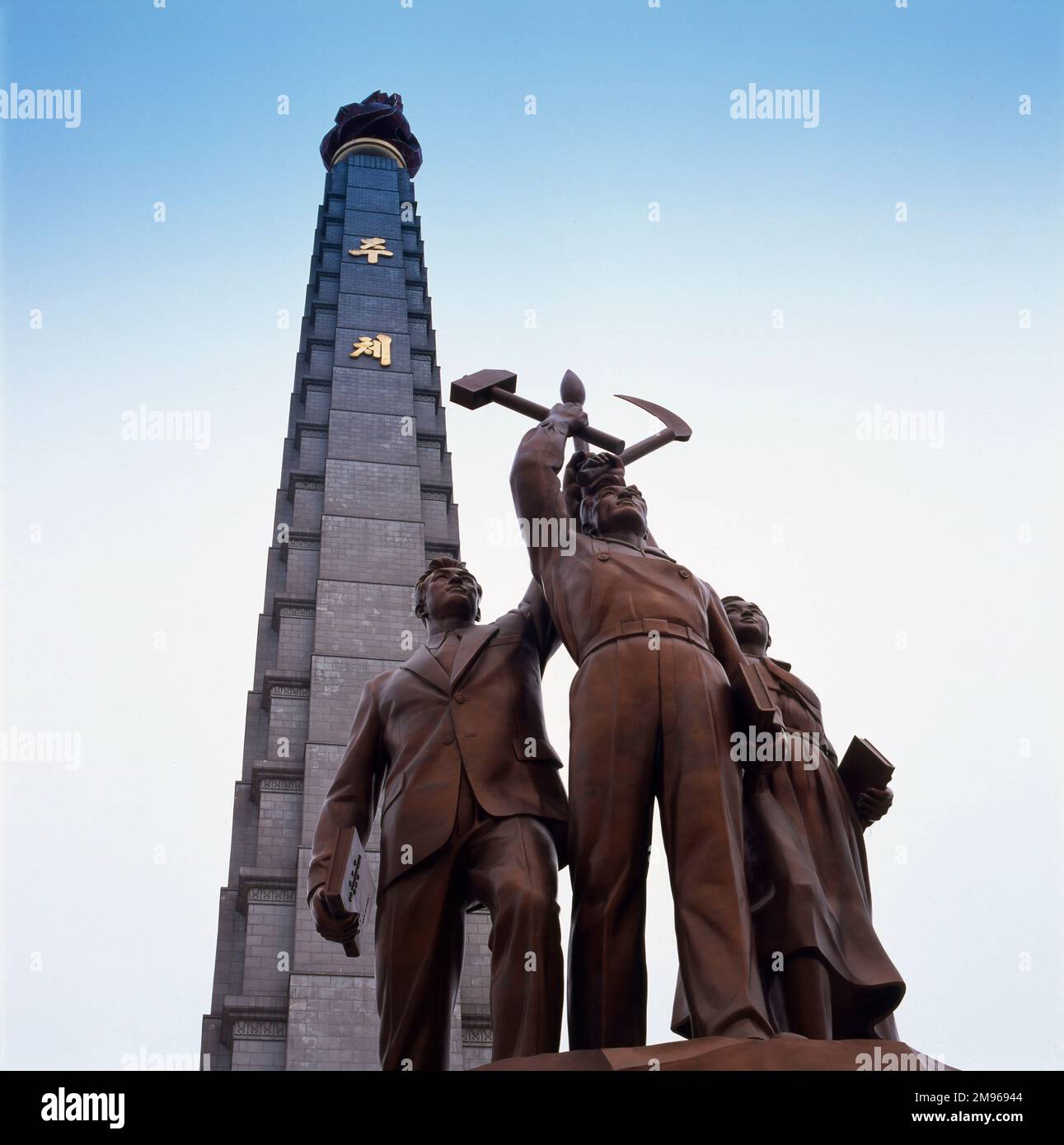 A statue depicting three members of the proletariat, one with a hammer, one with a sickle and one with a writing brush (an idealised worker, a peasant and a 'working intellectual'), at the foot of the Tower of the Juche Idea, on the eastern bank of the Taedong River in Pyongyang, capital of North Korea.   The tower is named after the principle of Juche, developed by Kim Il Sung (1912-1994) as a blend of autarchy, self-reliance, isolationism, Korean traditionalism, and Marxism-Leninism.  It was completed in 1982, to commemorate Kim Il Sung's 70th birthday, and is 170 metres high.  The associa Stock Photo