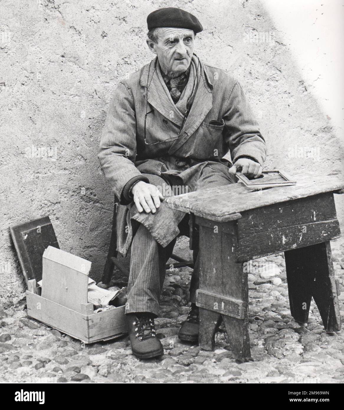 An old man (possibly Spanish) working on the street.  He wears a scruffy overcoat, a beret, and his glasses on a string round his neck.  He has a small wooden work bench and a box of materials.  Perhaps he is a picture framer. Stock Photo