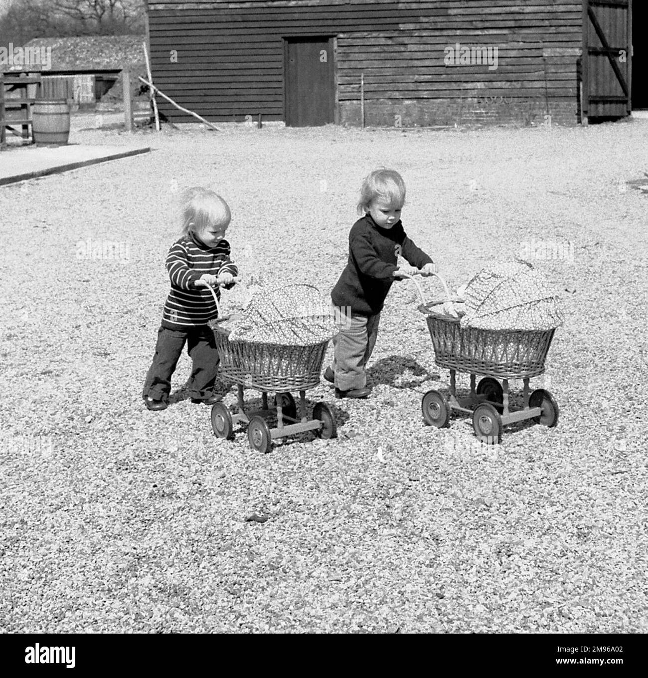 Two toddlers wheeling carry cots across a stretch of gravel. Stock Photo