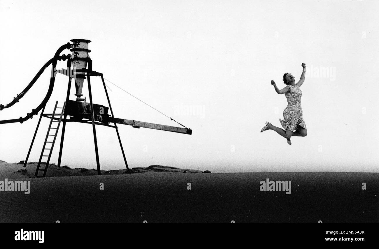 A middle aged woman in a flowery dress jumping for joy, near a gravel pit. What has made her so