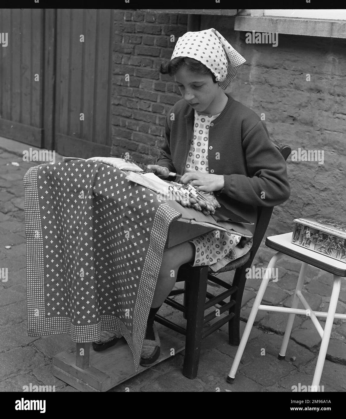 A young girl making lace on the pavement with a large number of bobbins