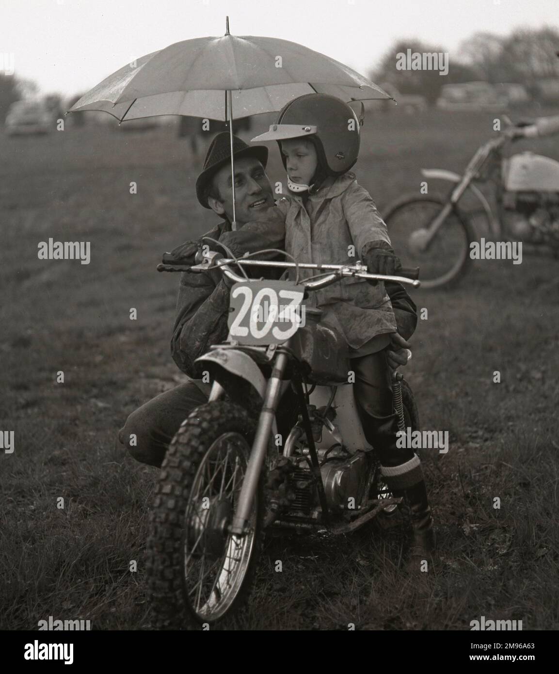 A man gives his son a bit of advice under an umbrella before the boy sets off on his junior sized motorcycle. Stock Photo