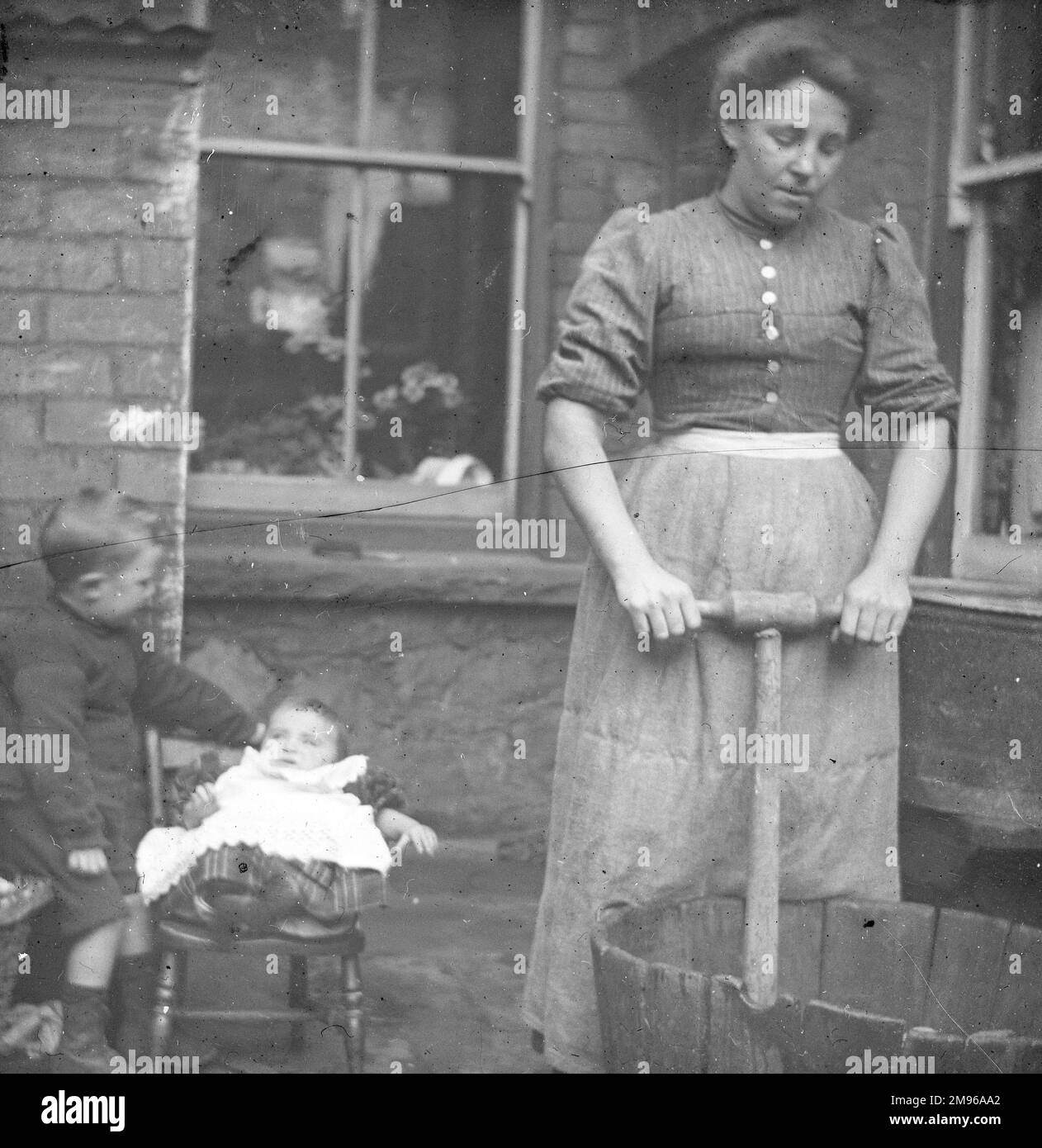 A woman using a dolly tub to do the weekly wash, in a back yard in South Wales, with two children on the left.  W E Jones recalled: 'Mrs George used to come and wash once a week for my mother and there you can see the old dolly tub.  There's two of my brothers, Lyndon the youngest one is in the chair.' Stock Photo