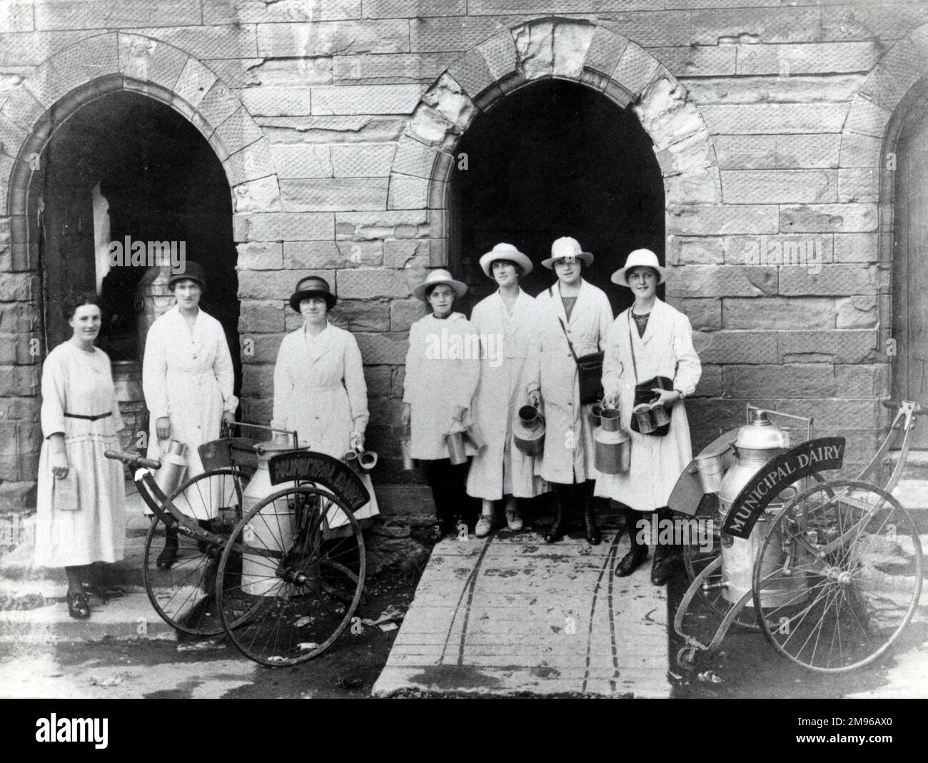 A group of seven women working for the Municipal Dairy pose outside the dairy building, somewhere in South Wales.  Two churn wagons, each one holding a large milk churn, can be seen in front of them.  The women hold smaller churns in their hands. Stock Photo