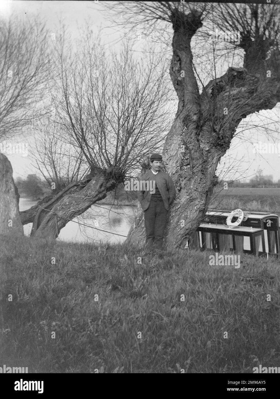 An Edwardian man poses by a tree on a canal bank, with a canal boat