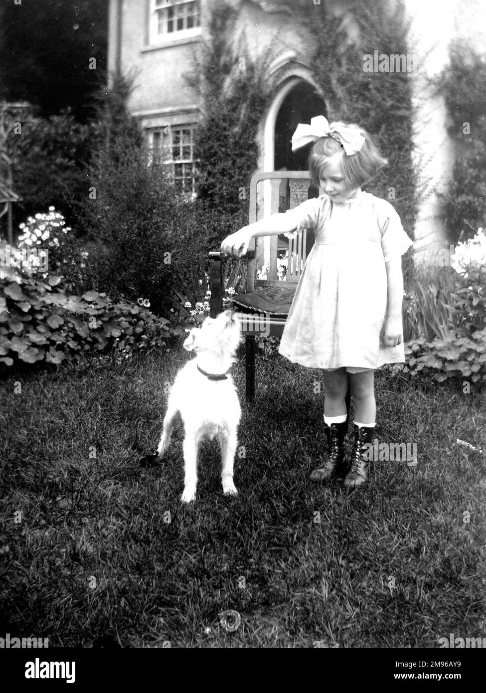 A little Edwardian girl in a garden with a small wire-haired terrier dog.  She is holding something in her hand for the dog to jump up and catch hold of. Stock Photo