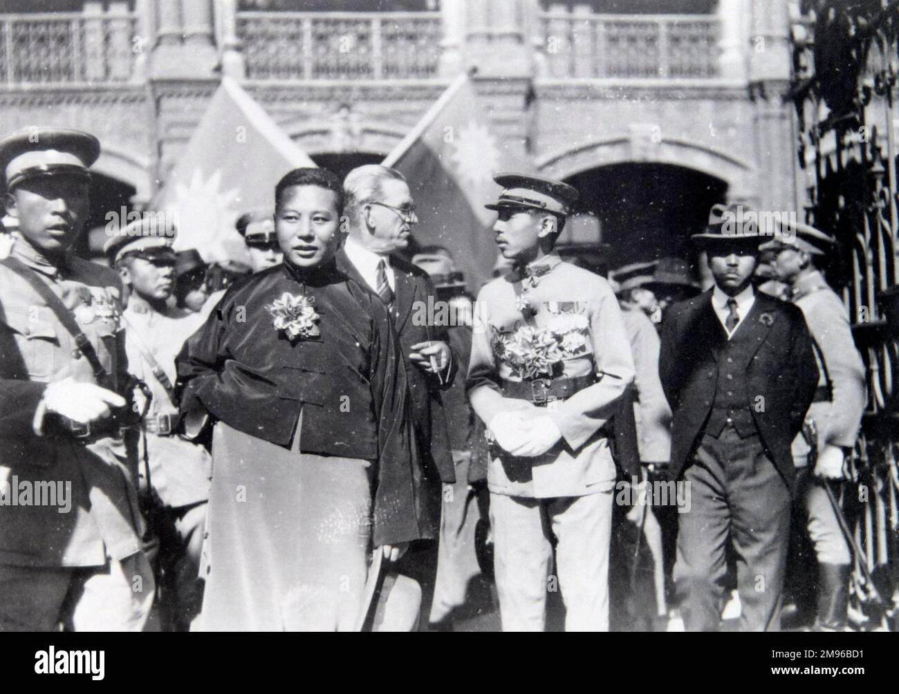 The local military chief, with others, some in uniform, others in civilian dress, somewhere in the Far East.  There is one Western man, perhaps a diplomat, talking to the chief.  The flags in the background seem to be those of the Republic of China (Taiwan). Stock Photo