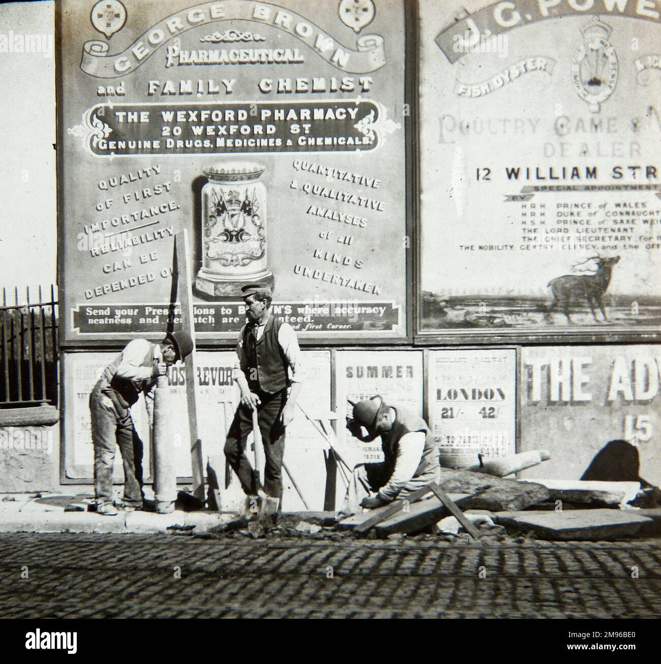 Three men busy doing street repairs in Dublin, Ireland.  The man on the left is laying a paving slab.  There are some rather ornate advertisements on the hoarding behind them, one for a family chemist, and one for a poultry, game and fish dealer. Stock Photo