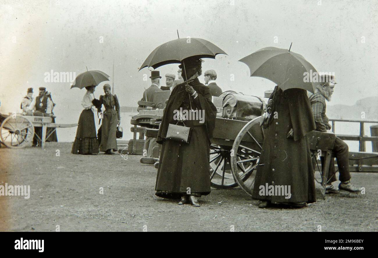 A group of Edwardian or late Victorian passengers waiting to board a boat, with a porter sitting on a luggage cart (right), and another porter with a cart (left).  It appears to be raining, as three umbrellas are up. Stock Photo