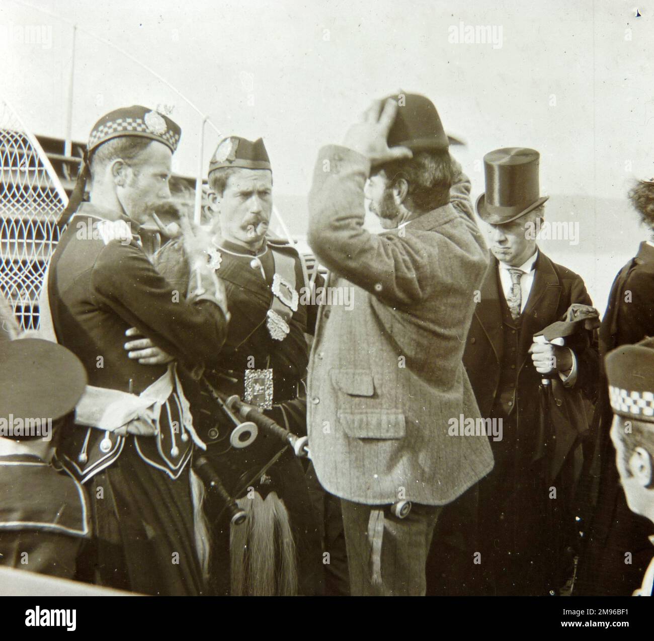 A group of passengers waiting to board a boat.  There are two Scottish pipers in traditional costume on the left, a man in a bowler hat in the middle, and a man in a top hat on the right. Stock Photo