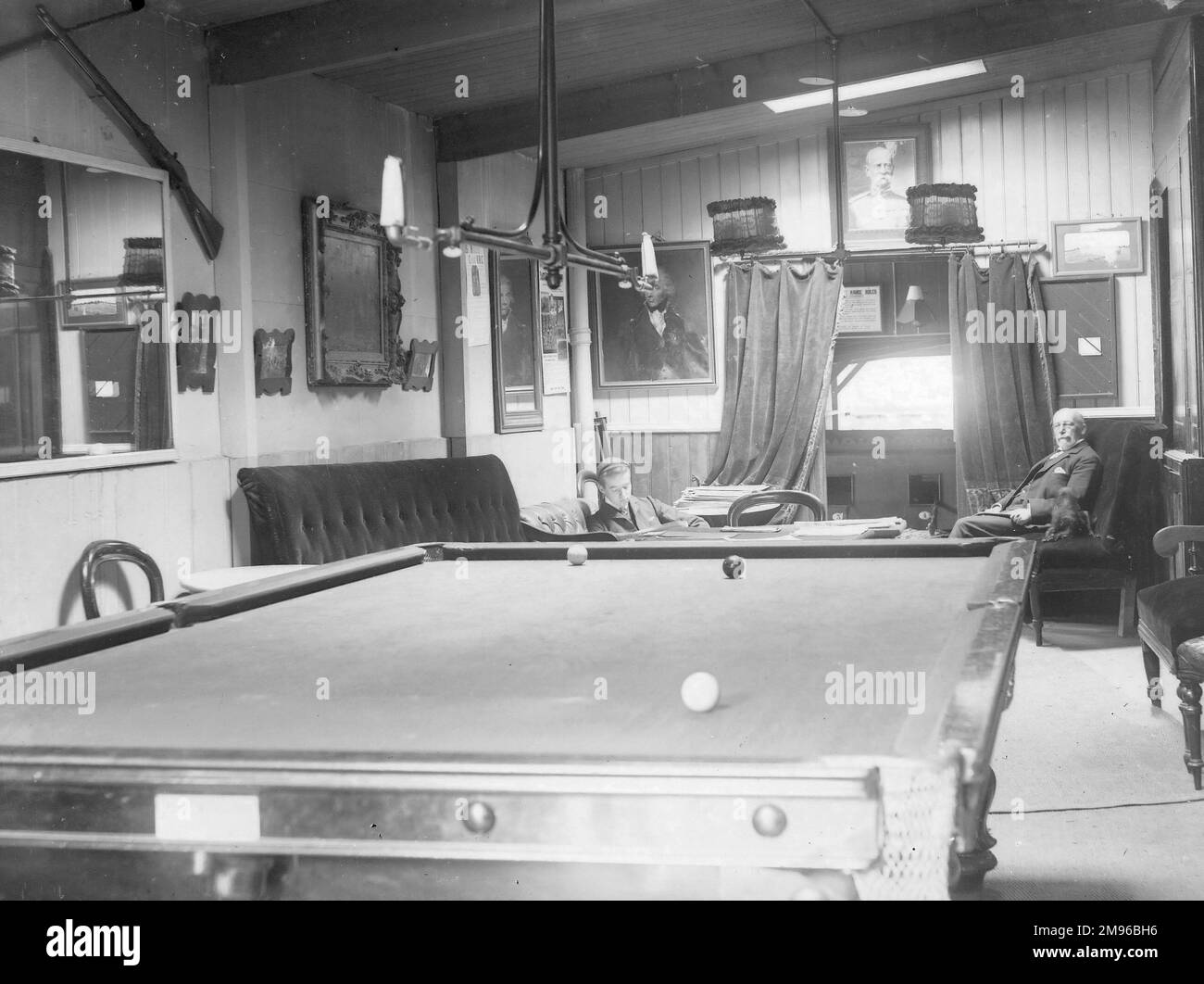 View inside a snooker room, with two players.  The snooker table is in the foreground, with three balls on it.  The players are both seated in the background. Stock Photo