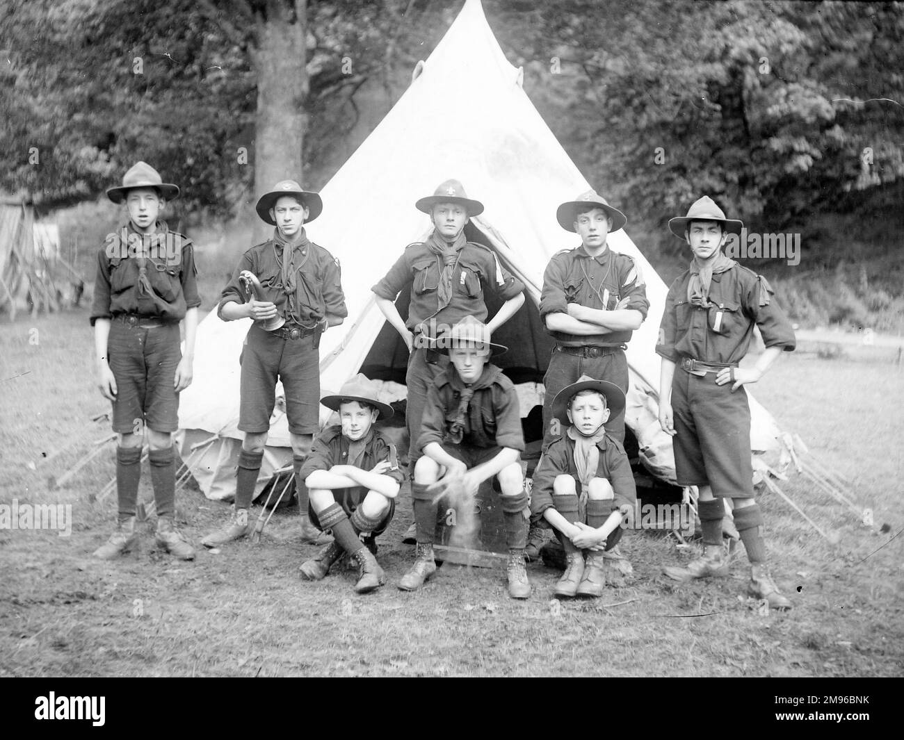 A group of eight boy scouts pose for their photo in a field, in front of a tent.  One of them is holding a bugle, no doubt used to wake them all up in the morning. Stock Photo