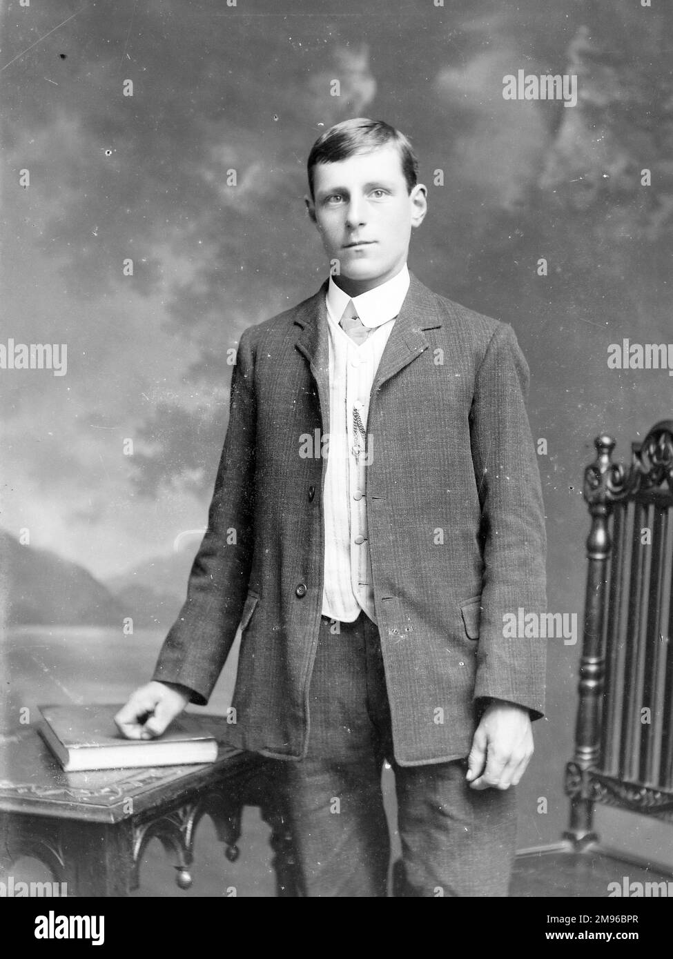 A young man poses in the photographer's studio, Mid Wales, with a book on a table on the left, and a carved chair on the right.  There is a painted backdrop behind him of a lake and mountains. Stock Photo