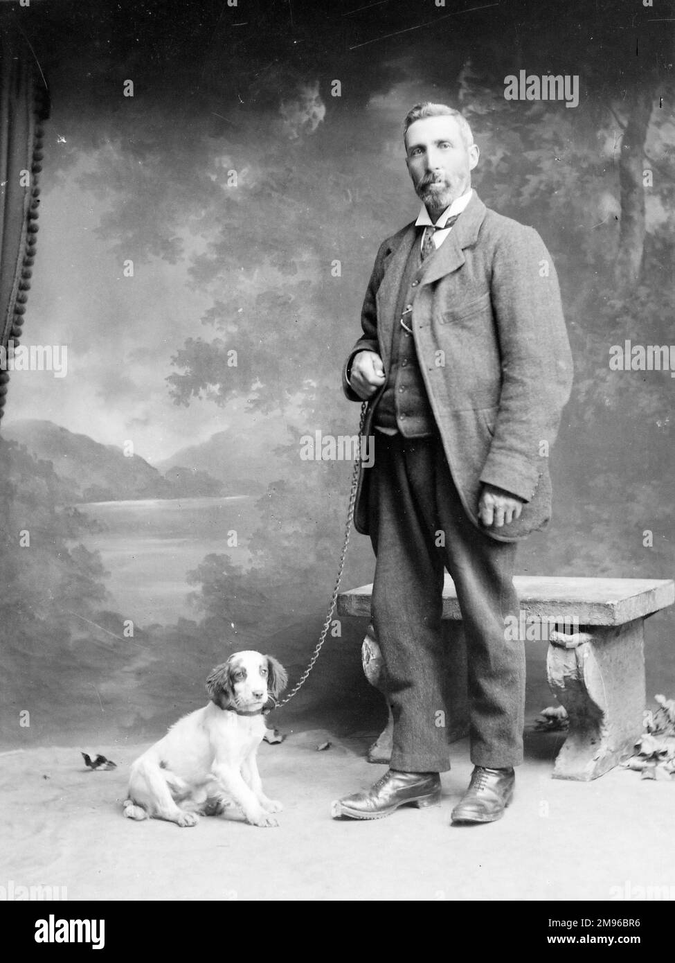 A middle aged man poses in the photographer's studio, Mid Wales, with a spaniel puppy on a chain.  There is a stone bench behind him, and a painted backdrop of a lake and mountains. Stock Photo