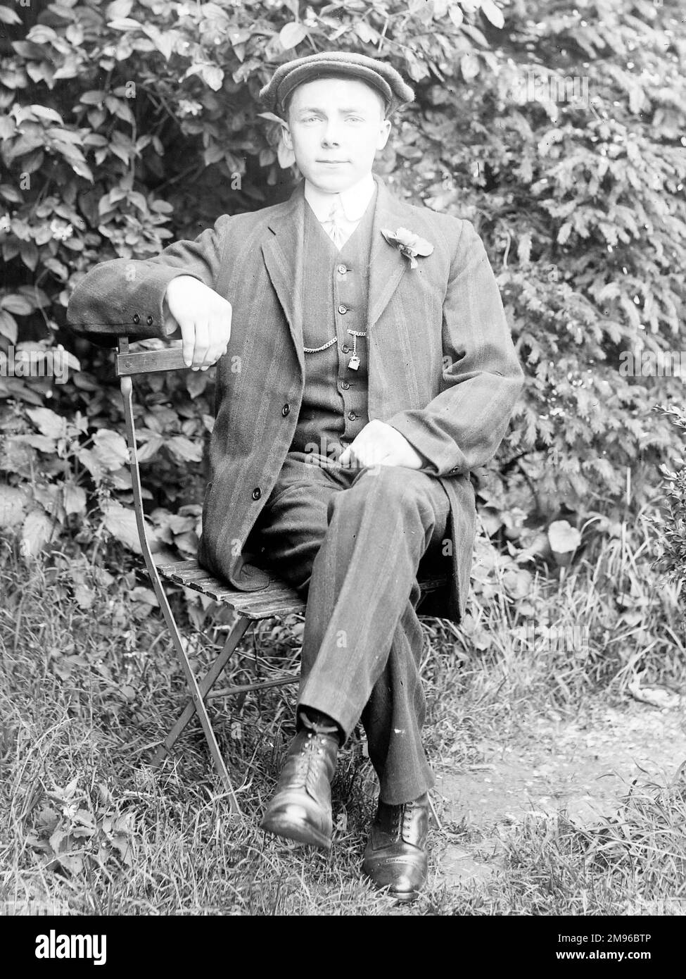 A dapper young man sitting on a folding steel chair in a garden, Mid Wales. He is wearing a