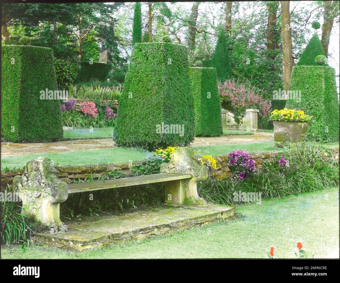 View of the gardens at Sedgewick Park House, near Horsham, West Sussex.  A stone bench can be seen in the foreground, with carefully shaped bushes behind, as well as grass, flowers and trees. Stock Photo