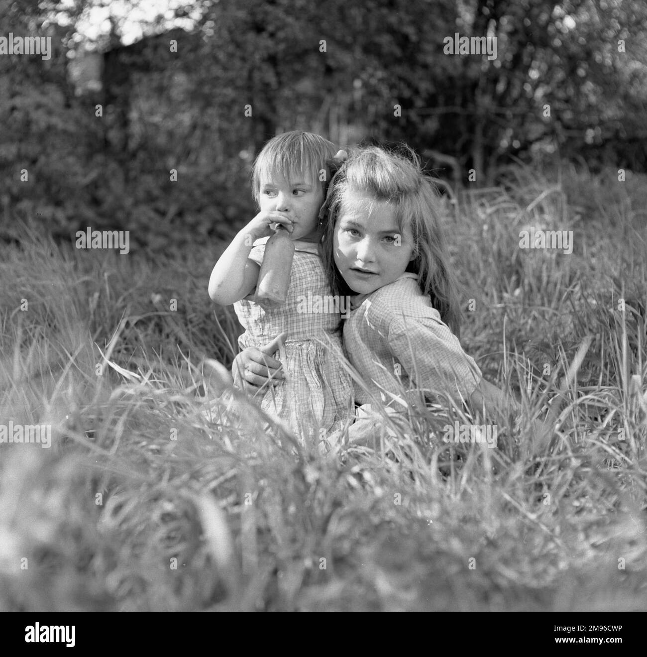 Two gipsy girls sitting in the long grass in a field.  The younger girl is holding a feeding bottle to her mouth. Stock Photo