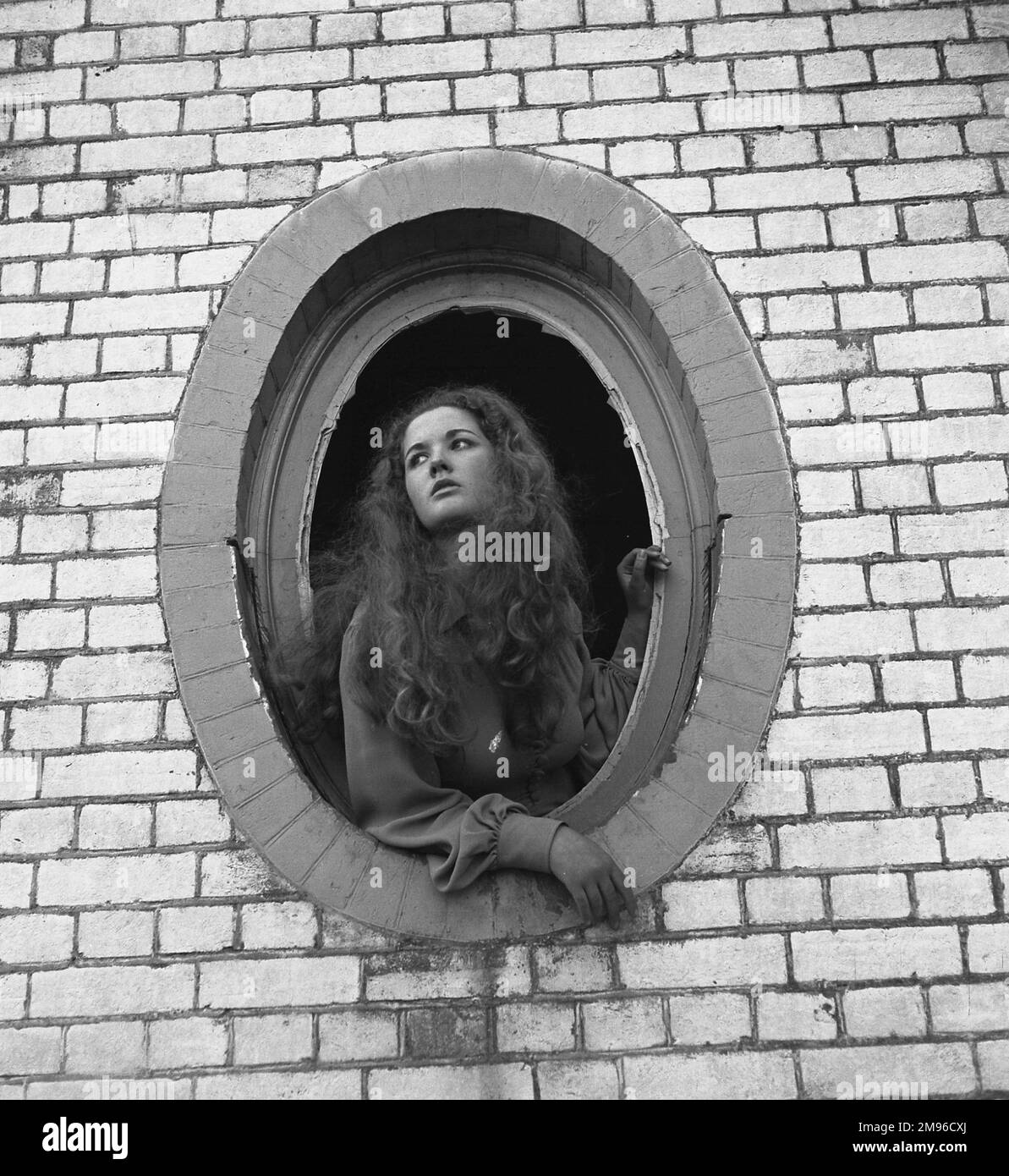 A young woman leans out of an oval window with broken glass, part of a derelict house. Stock Photo