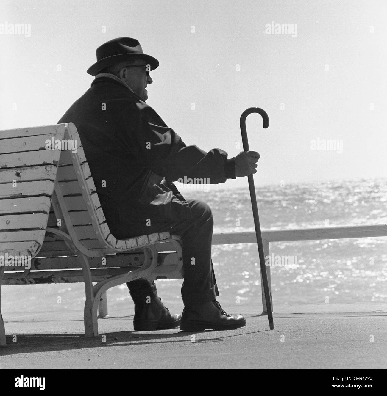 An old man with a walking stick, sitting on a bench looking out to sea. Stock Photo