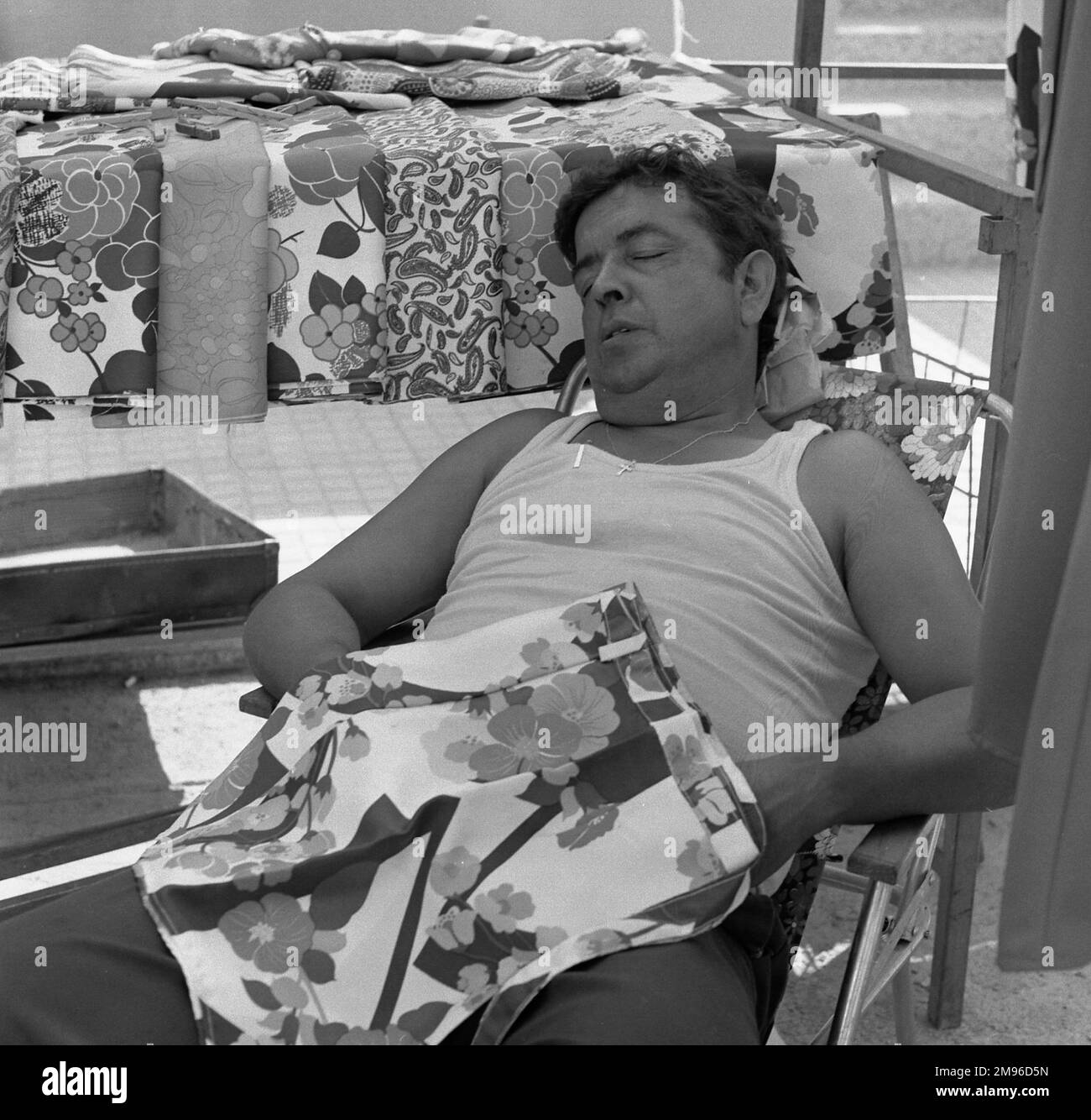 A man in a white vest sleeps in a chair, partially covered by some flowery patterned material.  Similar material covers a nearby table, possibly belonging to a street market. Stock Photo