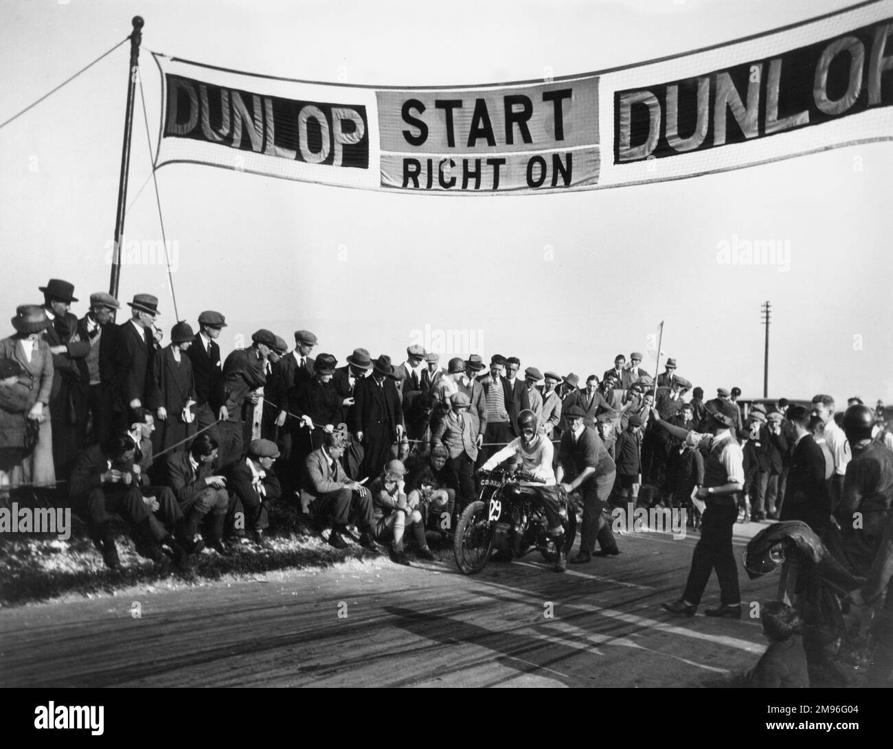 Crowds gather around a racetrack in Lewes, Sussex as man on a motorcycle prepares to be pushed off to begin the race, under a start banner indicating the sponsor, Dunlop. The Lewes Speed Trials were organised by Brighton & Hove Motor Cycle and Light Car Club. Stock Photo