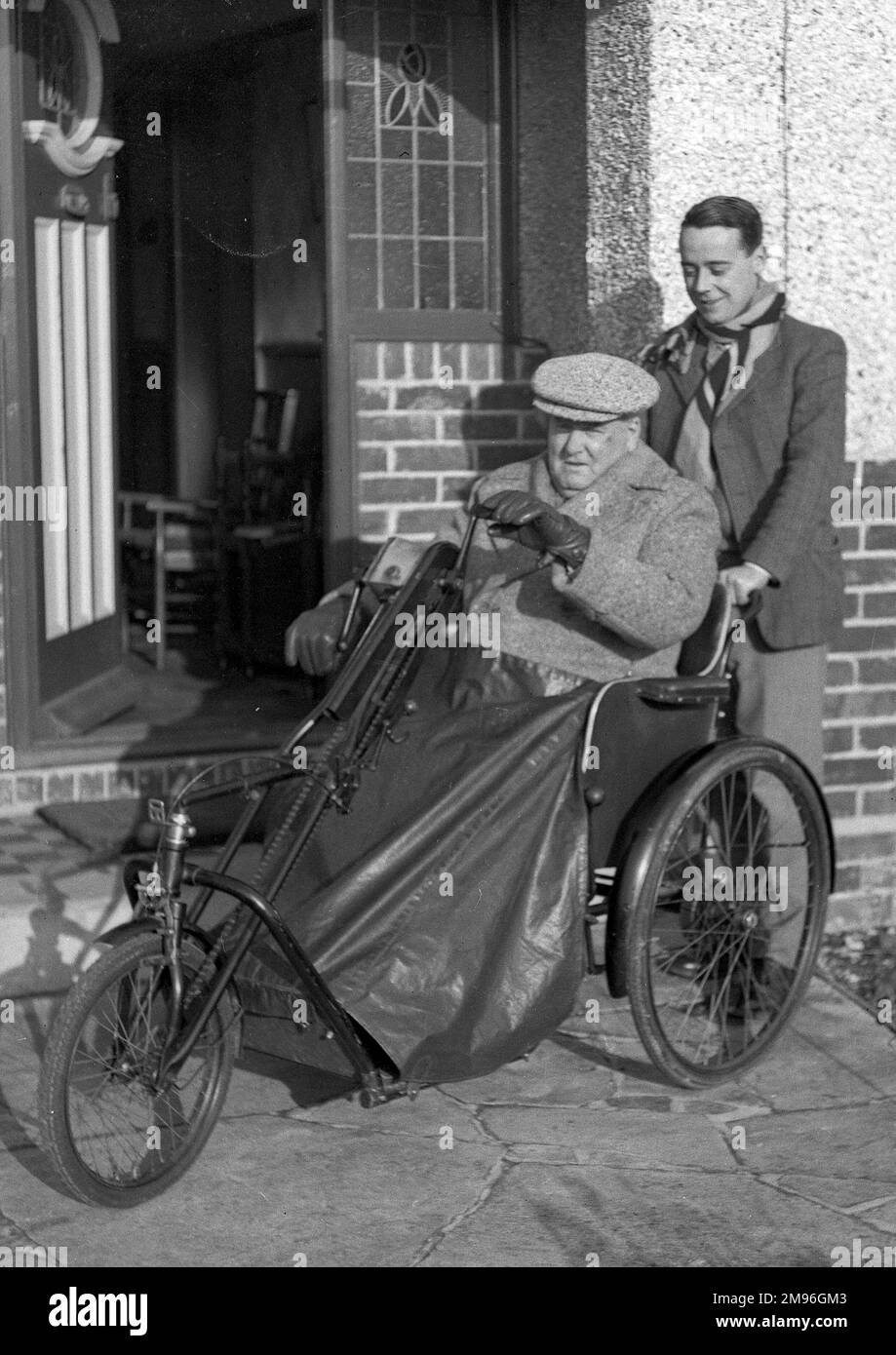 An elderly man in a bath chair at his front door, with a young man about to take him for an outing. Stock Photo