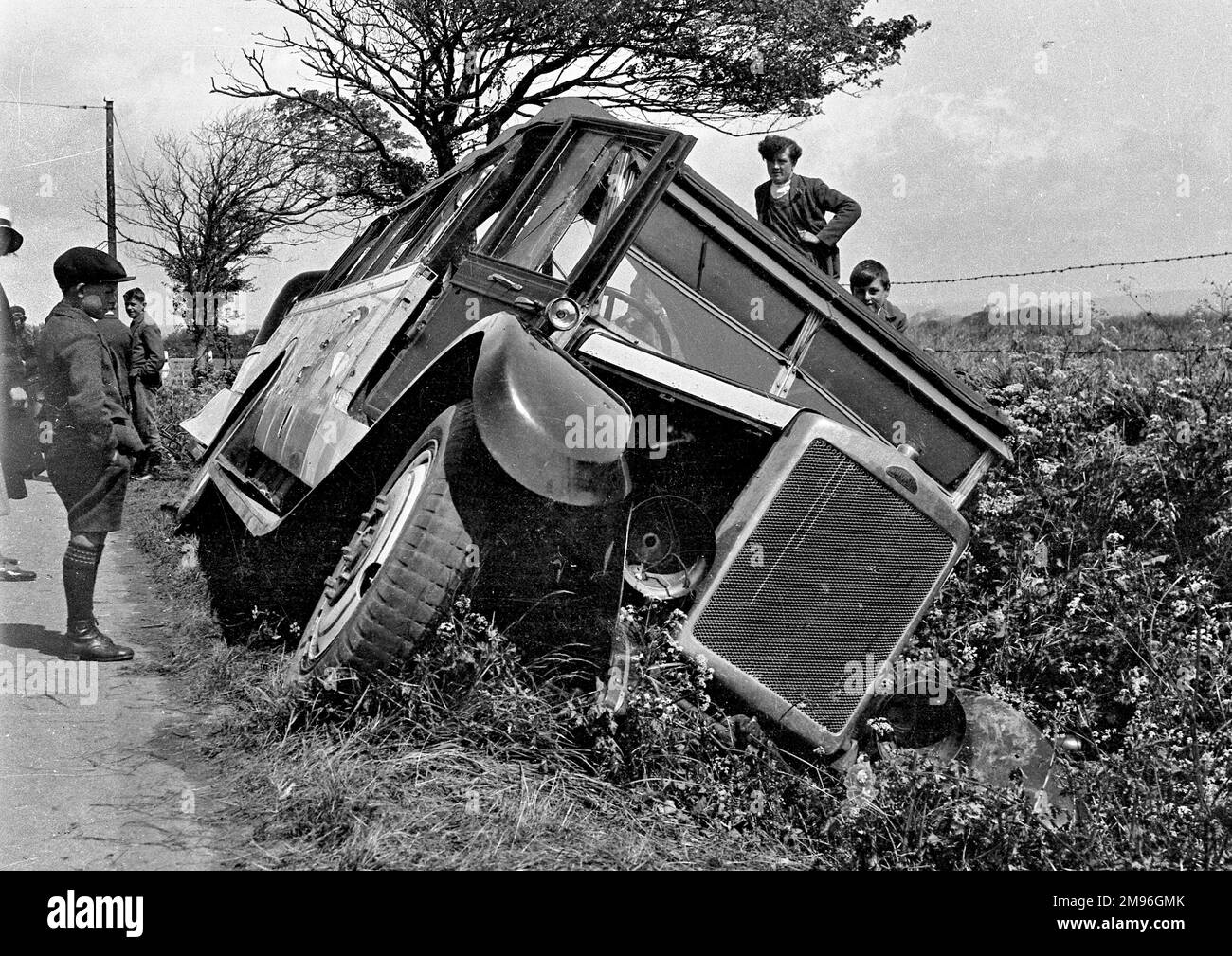 A bus which has somehow fallen into a ditch.  A group of boys stand around, wondering how to get it out again. Stock Photo