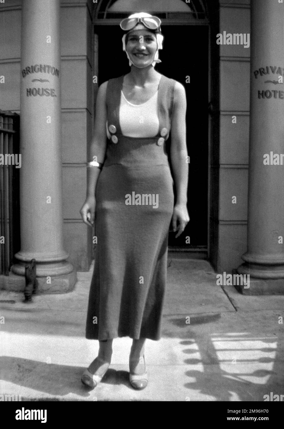 A smiling young woman wearing a woollen dress and goggles on her head stands outside Brighton House, a private hotel. Stock Photo