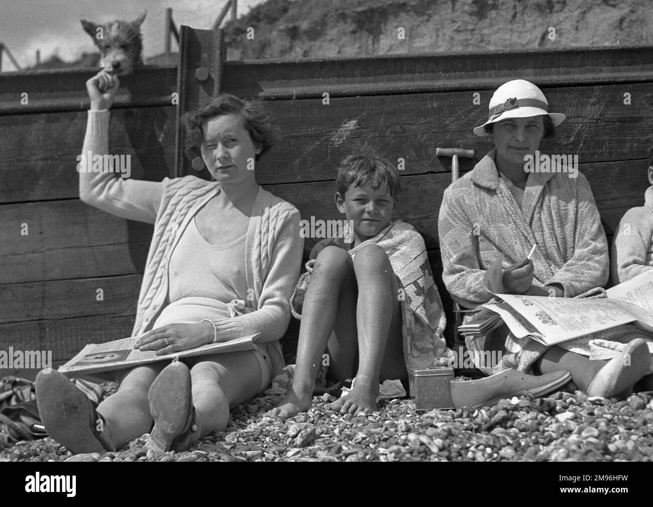 People on holiday, leaning against a breaker on a pebbly beach.  The woman on the left is holding up some food for a little dog. Stock Photo