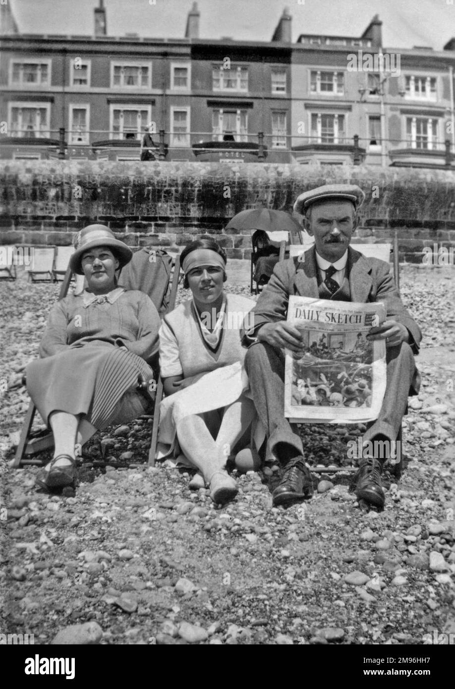 Three holidaymakers, two women and a man, sitting on the pebbly beach at Hastings, Sussex.  He is reading the Daily Sketch newspaper.  A terrace of hotels can be seen in the background. Stock Photo